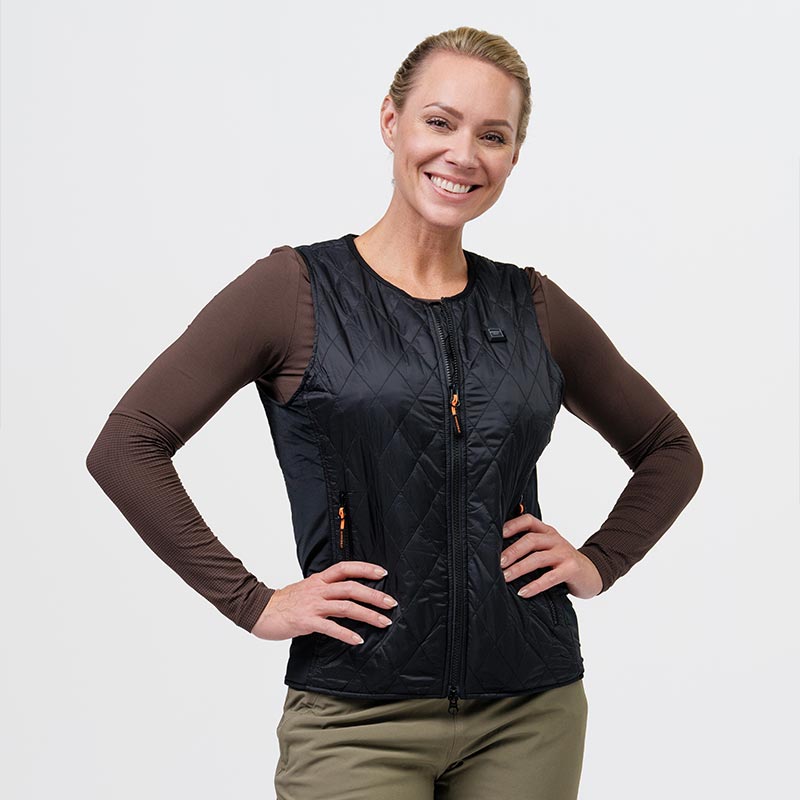 En kvinde bærer en sort, quiltet varmevest fra NordicHeat, står med hænderne i siden og smiler, iført en brun langærmet undertrøje. Baggrunden er lys.
