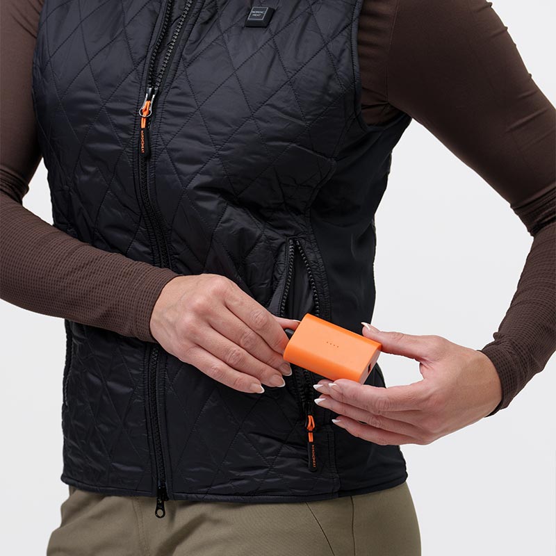 En person indsætter et orange batteri i lommen på en sort, quiltet jakke fra NordicHeat. Jakken bæres over en brun trøje.