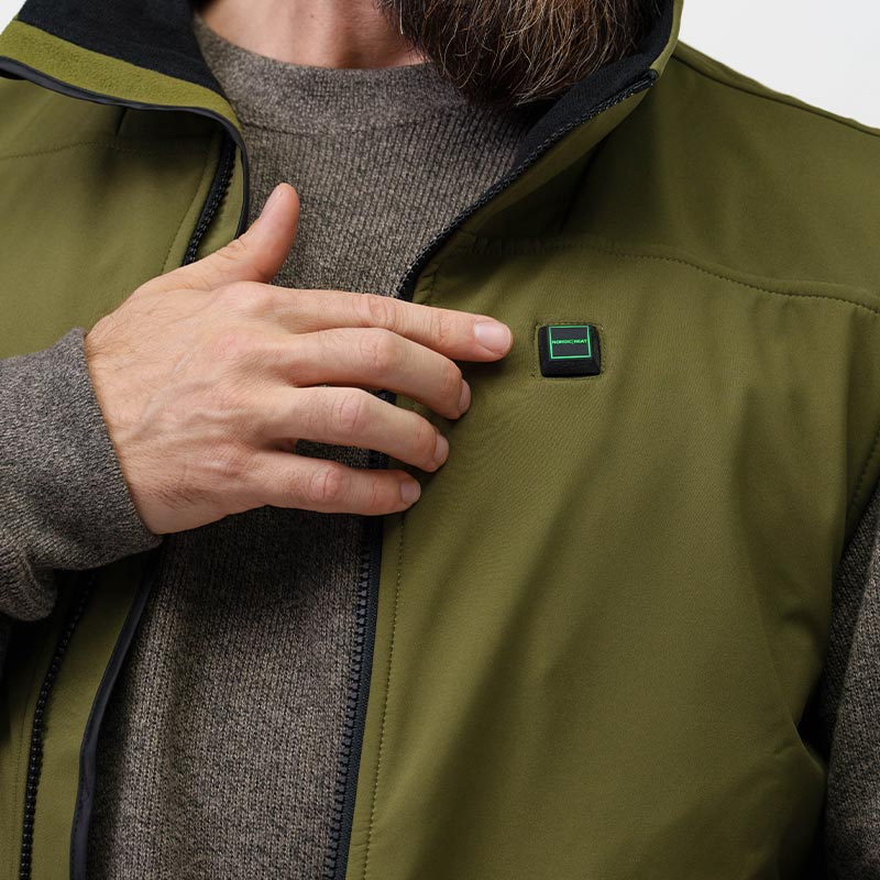En person iført en olivengrøn jakke rører ved en lille firkantet knap på brystet, hvor der står "NORDIC HEAT." Jakken bæres over en grå sweater.