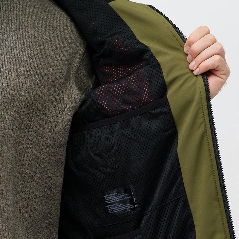 En hånd holder en åbnet jakke i olivengrøn, hvor det ses, at jakken har et sort, perforeret for med røde varmeelementer. Der er en etikette med tekst nederst.