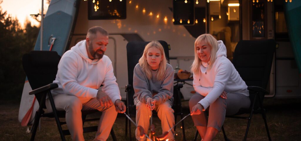Tre personer ristet skumfiduser over et bål foran en campingvogn i komfortabelt varmetøj. Velegnet til aktive udendørsoplevelser. Ingen synlig tekst.