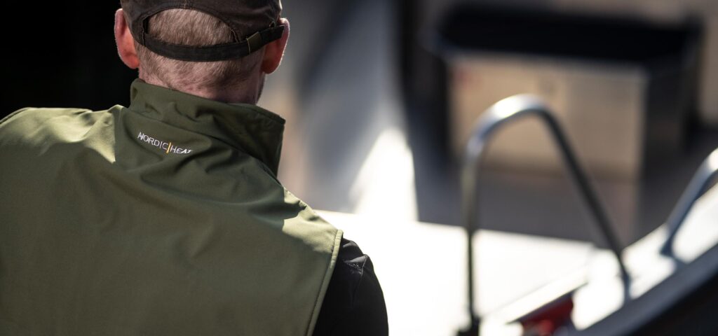 Person iført grøn NordicHeat jakke med tekst "NORDIC | HEAT" står ved paraplystativ i udendørs miljø, antyder moderne varmetøj til aktiv livsstil.